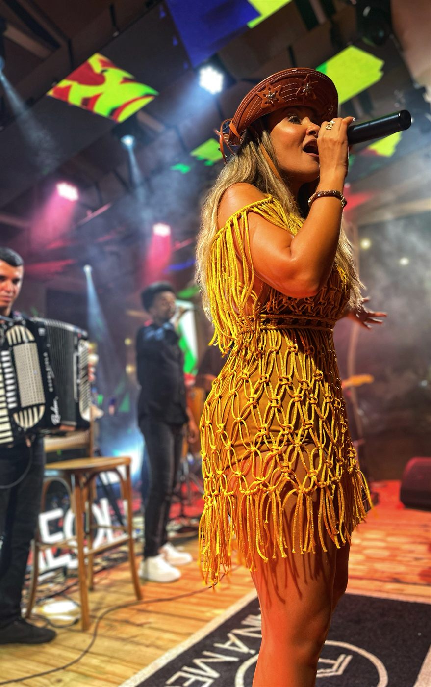 Tainá Meoli com figurino dourado e chapéu de couro, sanfoneiro ao fundo