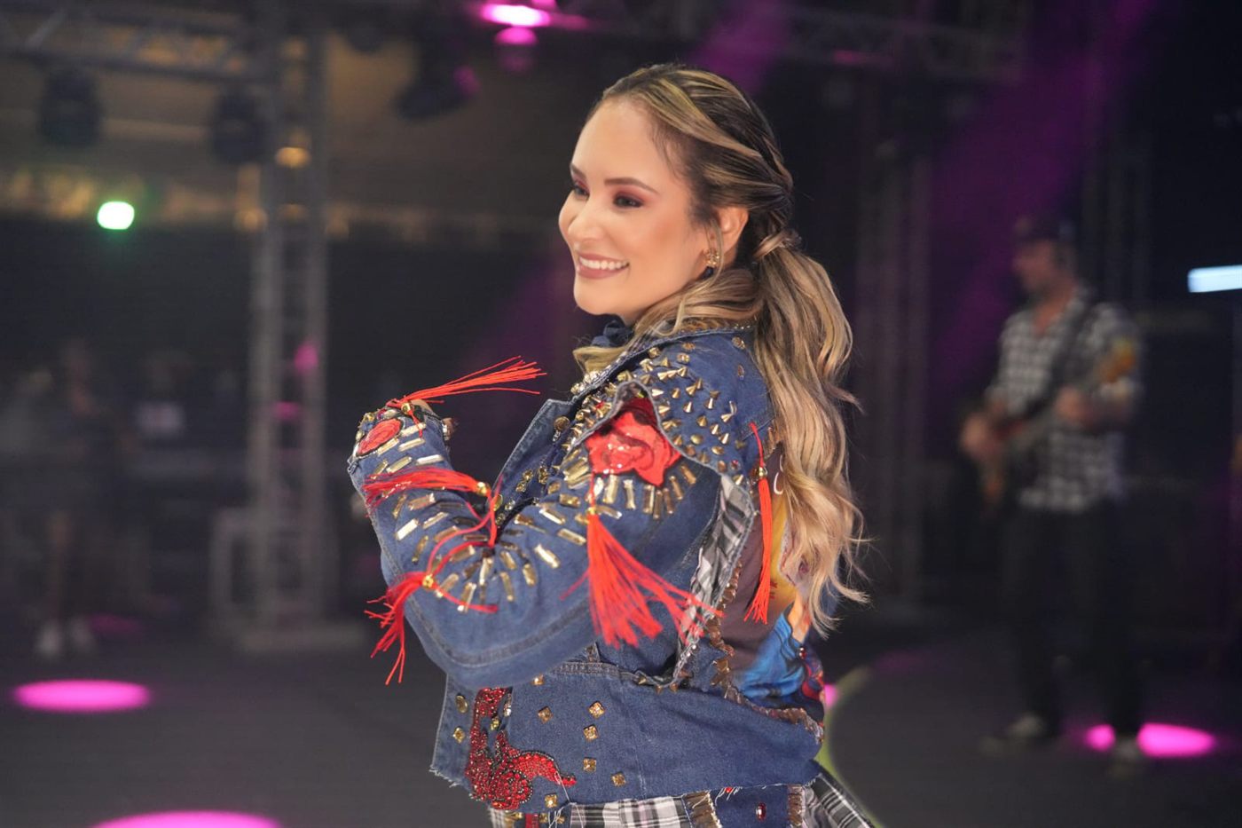 Tainá Meoli de perfil com jaqueta bordada, luzes rosas no palco