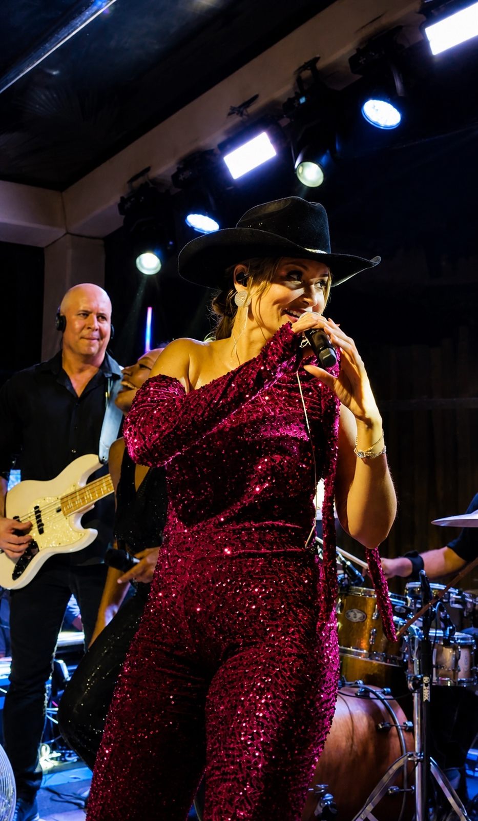 Tainá Meoli de macacão pink e chapéu preto cantando no palco